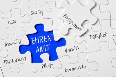 Ein Puzzle, bestehend aus mehreren weißen Puzzleteilen, die jeweils beschriftet sind. Es stehen dort die Worte: "Hilfe, sozial, freiwillig, Förderung, Pflege, Gemeinde, Verein, Tätigkeit. Ein letztes Puzzleteil muss noch in ein freies Feld eingesetzt werden. Das Teil ist mit dem Wort "Engagement" beschriftet. Auf dem freien Feld des Puzzles steht wiederum "Ehrenamt".