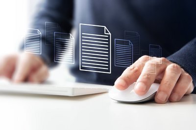 Eine Schreibtischoberfläche, auf der sich zwei Hände abstützen. In der linken Hand wird eine Computermaus gehalten. Die rechte Hand tippt etwas in eine Tastatur ein. Über den Händen schweben mehrere Dokumentensymbole