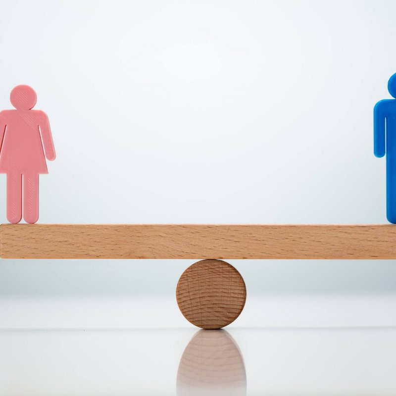 Eine Wippe, die sich im Gleichgewicht befindet. Auf der rechten Seite befindet sich der Umriss einer Frau und auf der linken Seite der Wippe der Umriss eines Mannes.