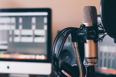 Ein Mikrophon in einem Tonstudio. Im Hintergrund stehen zwei Bildschirme, auf denen Tonaufnahmen abgebildet sind.